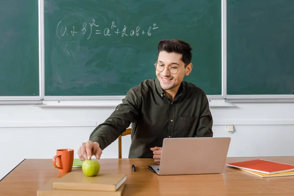 Insegnante maschio sorridente in occhiali seduto alla scrivania del computer e raggiungere per mela in classe — Foto stock