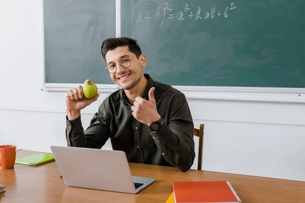 Maschio insegnante in occhiali guardando la fotocamera, tenendo mela e mostrando pollice su segno alla scrivania del computer in classe — Foto stock