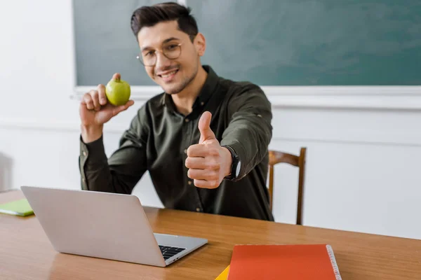 Sorridente insegnante di sesso maschile in bicchieri con mela in mano e mostrando il pollice al computer scrivania in classe — Foto stock