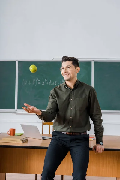 Улыбающийся учитель-мужчина бросает яблоко в классе с доской и столом на заднем плане — стоковое фото