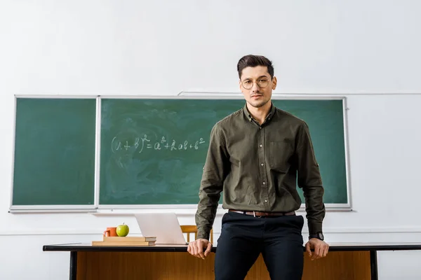 Profesor masculino serio en ropa formal mirando a la cámara y de pie cerca del escritorio en el aula - foto de stock