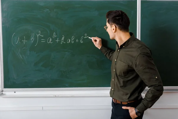 Maschio insegnante in abbigliamento formale scrittura equazione sulla lavagna in classe di matematica — Foto stock