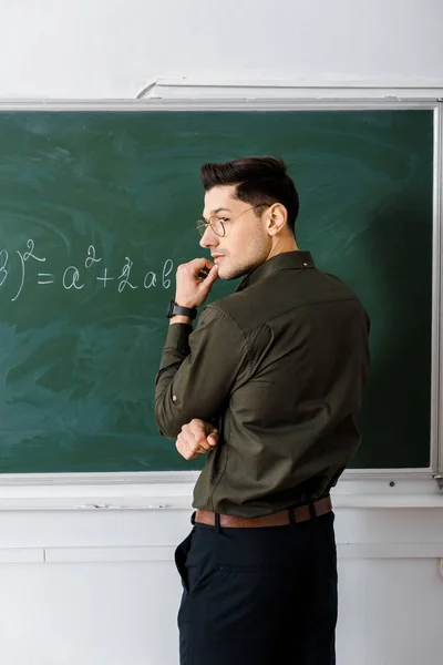Pensive male teacher touching chin and solving equation on chalkboard in class — Stock Photo