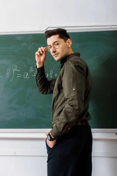 Красивый учитель мужского пола держит очки и смотрит на камеру в классе математики — стоковое фото