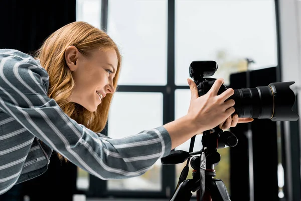 Visão lateral de sorrir jovem fotógrafa trabalhando com câmera fotográfica profissional em estúdio — Fotografia de Stock