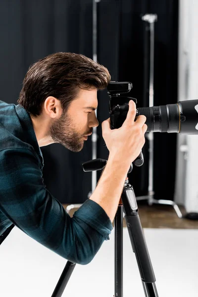 Vista lateral de joven fotógrafo guapo trabajando con cámara fotográfica profesional en el estudio - foto de stock