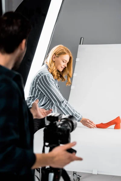 Cropped shot of photographer looking at smiling female colleague arranging shoe in studio — Stock Photo