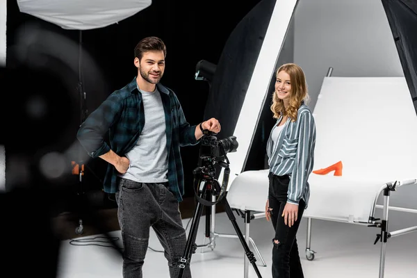 Heureux jeunes photographes debout près de matériel photo professionnel et souriant à la caméra en studio — Photo de stock