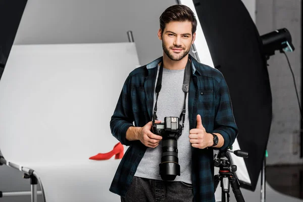 Красивый улыбающийся молодой фотограф держит камеру и показывает большой палец в студии — стоковое фото