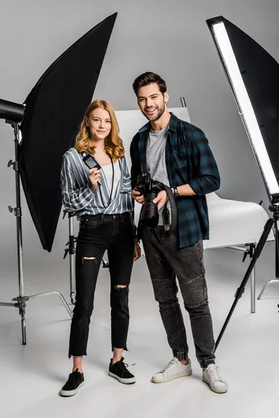 Happy young photographers with professional equipment standing together and smiling at camera in studio — Stock Photo