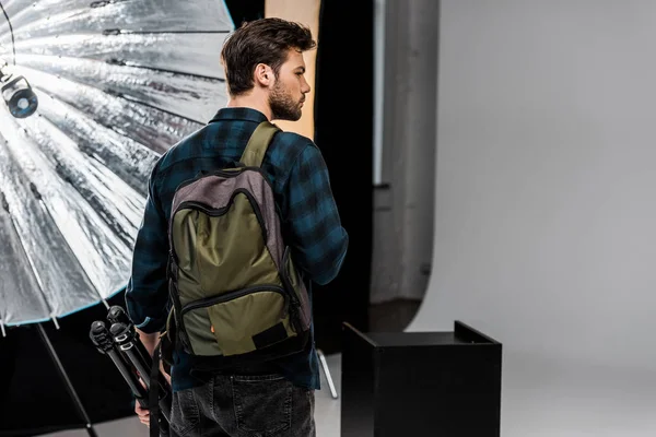 Vue arrière du jeune homme avec sac à dos tenant équipement professionnel et regardant loin dans le studio de photo — Photo de stock