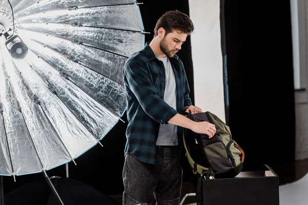 Молодой фотограф упаковывает профессиональное фотооборудование в рюкзак — стоковое фото