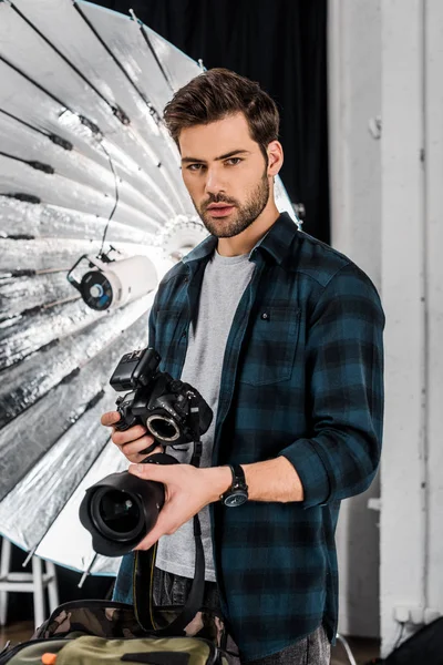Bonito profissional jovem fotógrafo segurando câmera e lente em estúdio — Fotografia de Stock