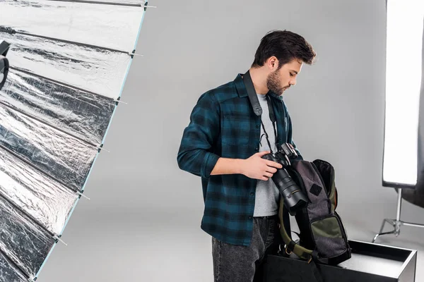 Fotógrafo joven profesional sosteniendo la cámara y la mochila con equipo fotográfico en el estudio - foto de stock