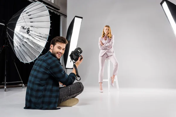 Fotografo seduto e sorridente alla macchina fotografica mentre bella modella in posa in studio — Foto stock