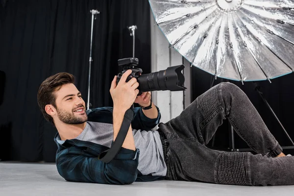 Jeune photographe souriant couché et utilisant un appareil photo en studio — Photo de stock