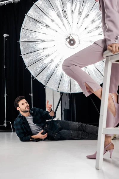 Sonriente joven fotógrafo con cámara tumbada y trabajando con modelo elegante en el estudio - foto de stock