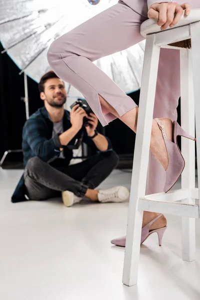 Recortado disparo de fotógrafo sonriente sentado y fotografiando elegante modelo femenino en el estudio - foto de stock