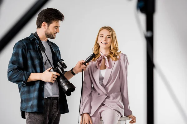 Sorrindo jovem fotógrafo segurando câmera e medidor de luz enquanto trabalhava com modelo em estúdio — Fotografia de Stock