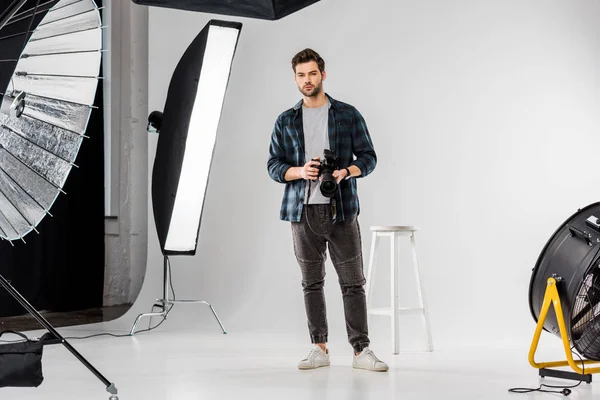 Bonito jovem fotógrafo segurando câmera profissional e olhando para a câmera no estúdio de fotos — Fotografia de Stock