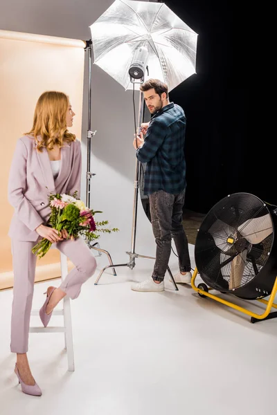 Young photographer working with lighting equipment and looking at beautiful female model with flowers in photo studio — Stock Photo