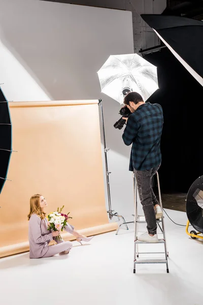 Fotógrafo de pie en la escalera de paso y disparar hermosa chica con flores en el estudio - foto de stock