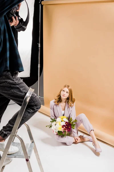 Schnappschuss von Fotograf, der auf Leiter steht und schönes Mädchen mit Blumen im Atelier fotografiert — Stockfoto