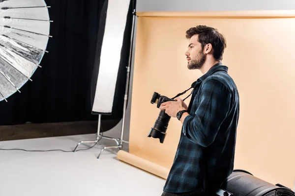 Vue latérale du beau jeune photographe tenant la caméra et regardant loin dans le studio de photo — Photo de stock