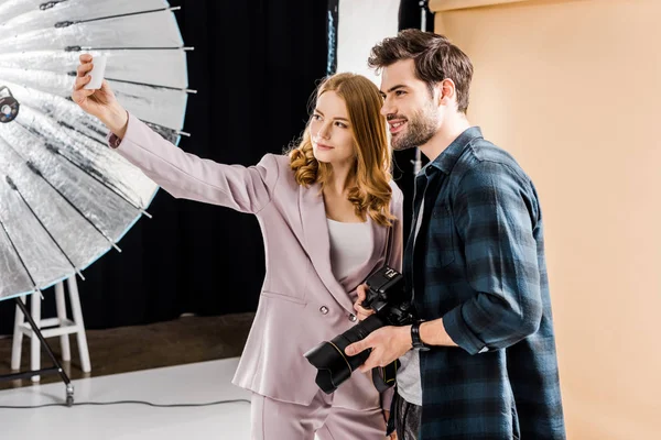 Joven fotógrafo guapo y hermosa modelo sonriente tomando selfie con teléfono inteligente en estudio de fotos - foto de stock