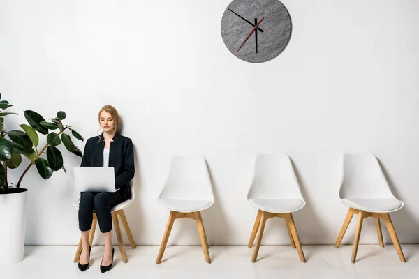 Joven mujer de negocios utilizando el ordenador portátil mientras está sentado en la silla y esperando en línea - foto de stock