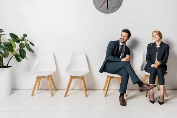 Jóvenes empresarios sonrientes sosteniendo vasos de papel y mirando hacia otro lado mientras esperan en sillas en fila - foto de stock
