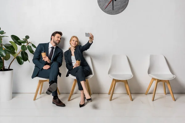 Gente de negocios sonriente tomando selfie con teléfono inteligente mientras se sienta en sillas en línea - foto de stock