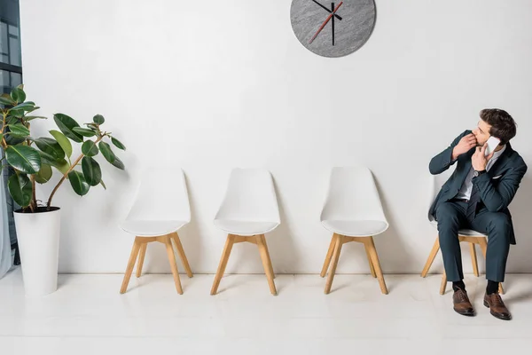 Nervioso joven hombre de negocios hablando por teléfono inteligente y mirando el reloj mientras está sentado en la silla en línea - foto de stock