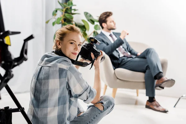 Jovem fotógrafa sorrindo para a câmera enquanto fotografa belo empresário em estúdio — Fotografia de Stock