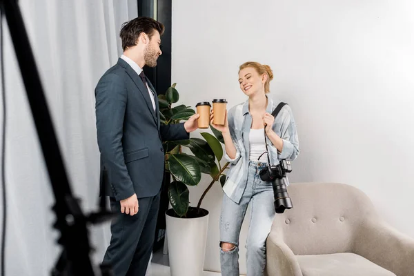 Photographe souriant et beau modèle tenant des tasses en papier et parlant en studio photo — Photo de stock