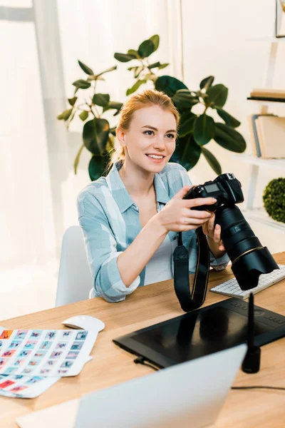 Hermosa sonriente joven fotógrafo sosteniendo la cámara mientras trabaja en la oficina - foto de stock
