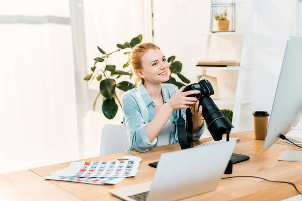 Hermosa sonrisa joven fotógrafa sosteniendo la cámara mientras trabaja en la oficina — Stock Photo