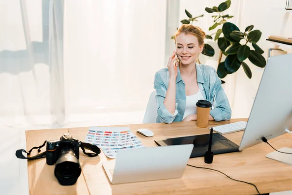 Lächelnde junge Frau spricht per Smartphone, während sie Fotos im Büro retuschiert — Stockfoto