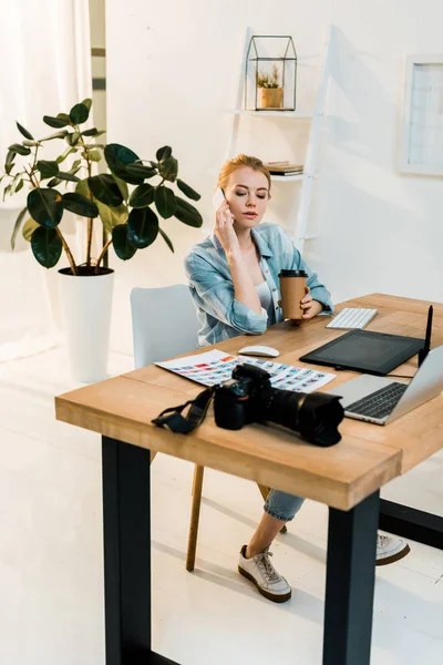 Hermoso joven retoucher sosteniendo café para ir y hablar por teléfono inteligente en el lugar de trabajo — Stock Photo