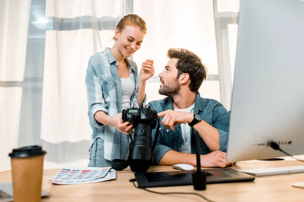 Fotógrafos jóvenes profesionales utilizando la cámara y sonriendo entre sí en el lugar de trabajo - foto de stock