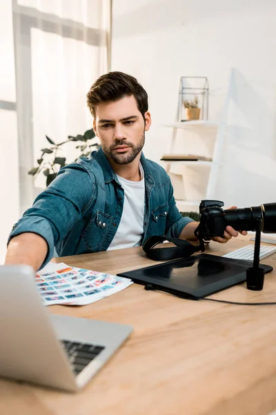 Joven fotógrafo guapo usando cámara y portátil en el lugar de trabajo - foto de stock