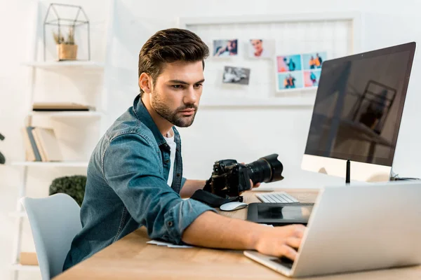 Bel giovane fotografo che tiene la fotocamera e utilizza il computer portatile in ufficio — Foto stock