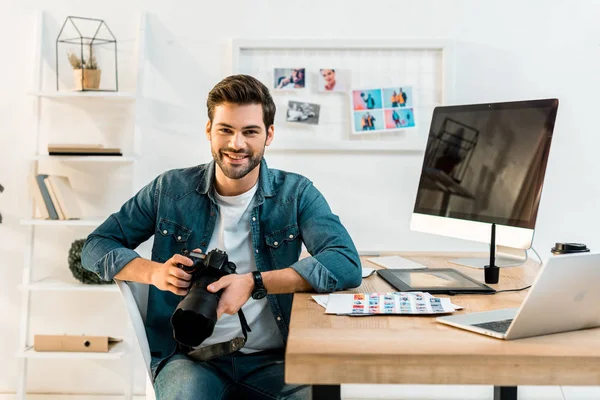 Bel giovane fotografo che tiene la fotocamera e sorride alla fotocamera in ufficio — Foto stock