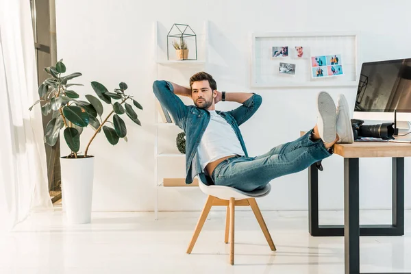 Hermoso joven retoucher sentado con las manos detrás de la cabeza y las piernas en el escritorio - foto de stock