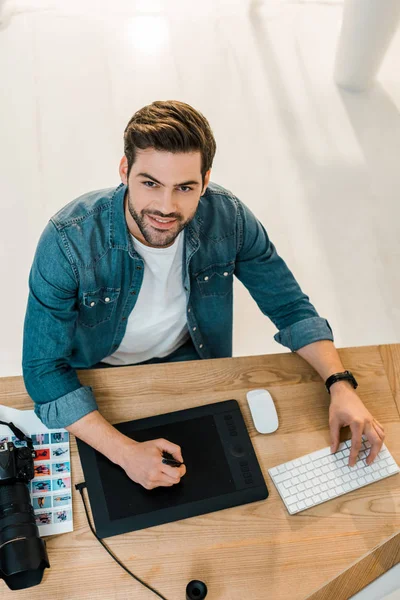 Vue grand angle de beau jeune retouche souriant à la caméra tout en travaillant dans le bureau — Photo de stock