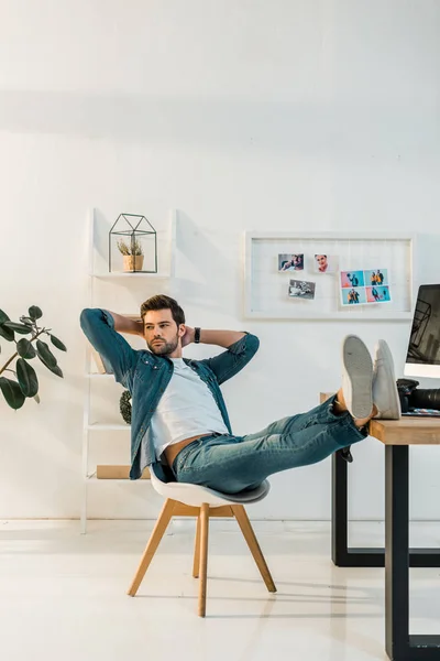 Relaxed young retoucher sitting with hands behind head and legs on work desk — Stock Photo