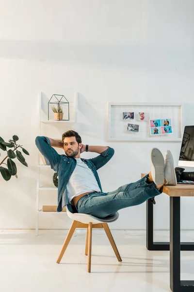 Relaxado jovem retoucher sentado com as mãos atrás da cabeça e pernas na mesa de trabalho — Fotografia de Stock