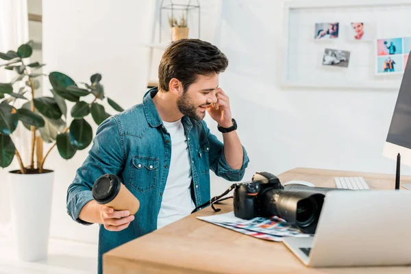 Smiling young retoucher holding coffee to go and talking by smartphone at workplace — Stock Photo