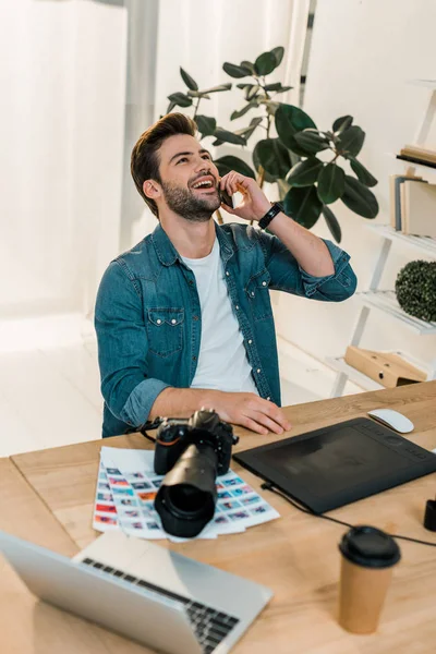 Cheerful young retoucher talking by smartphone and looking up at workplace — Stock Photo