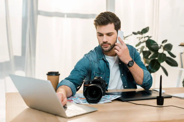 Молодий ретушер розмовляє по смартфону і використовує ноутбук на робочому місці — стокове фото